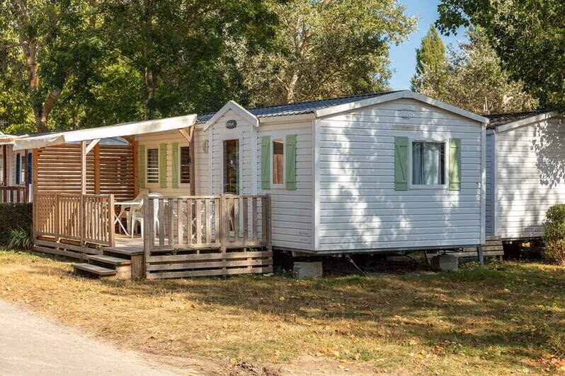 Witte stacaravan met groene luiken en een houten veranda met rieten afscheiding op Camping Bella Italia, tussen de loofbomen