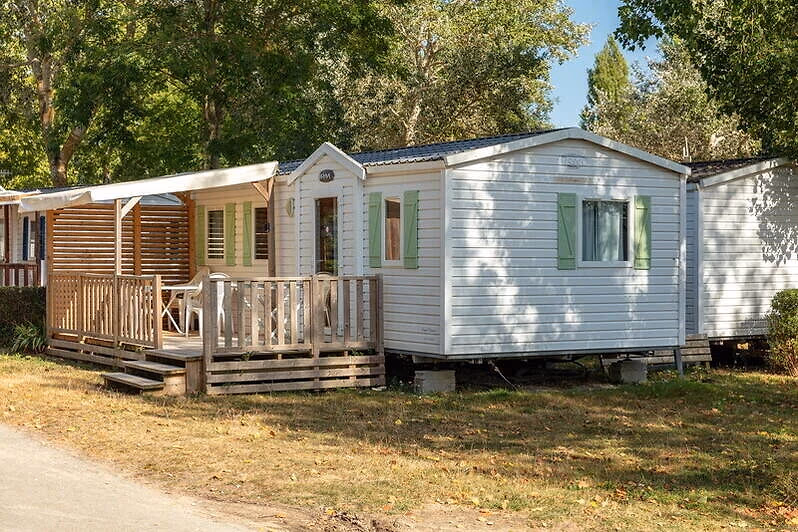 Witte stacaravan met groene luiken en een houten veranda met rieten afscheiding op Camping Bella Italia, tussen de loofbomen