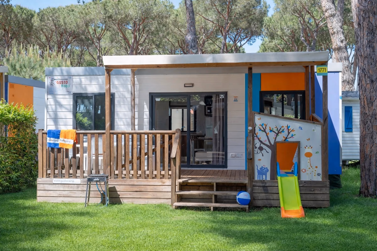 Stacaravan met een houten veranda op Camping Bella Italia, met een kindertekening op het schot, een glijbaan en een bal in het gras