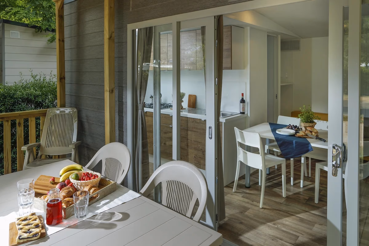Overdekt terras met een gedekte tafel en doorkijk door de vouwpui naar de keuken en eethoek van een stacaravan op Camping Bella Italia