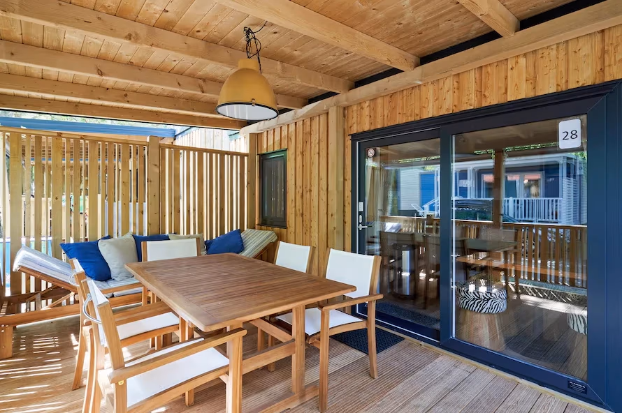 Houten terras onder een balkenkap met een teakhouten eettafel en een loungebank met blauwe kussens bij een stacaravan op Camping Bella Italia