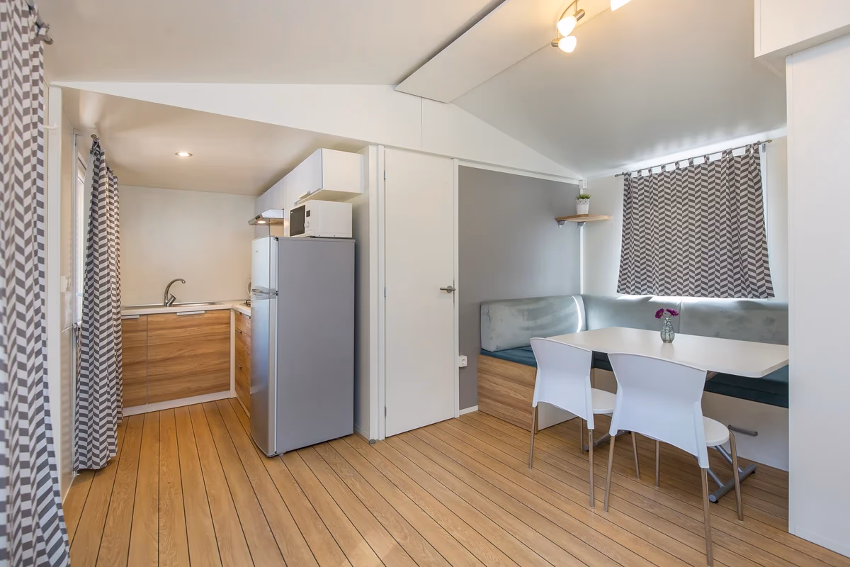 Woonruimte met een houten vloer, een kitchenette met koelkast en een eethoek met zitbank in een stacaravan op Camping Bella Italia
