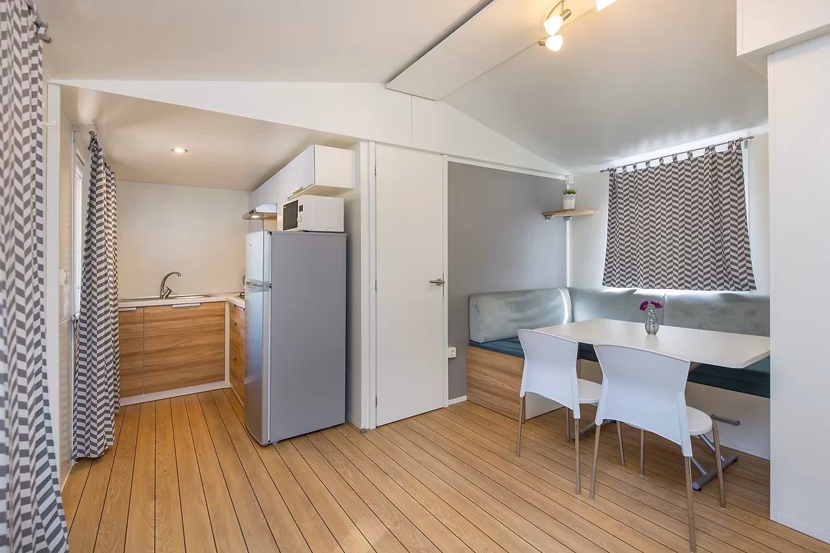 Woonruimte met een houten vloer, een kitchenette met koelkast en een eethoek met zitbank in een stacaravan op Camping Bella Italia