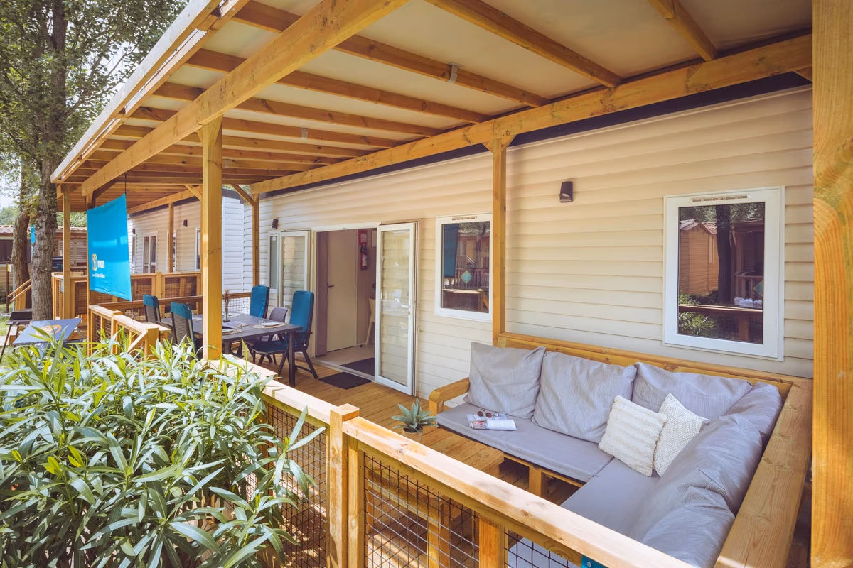 Overdekte veranda met een grijze hoekbank en een eettafel met blauwe stoelen bij een stacaravan, met oleanders langs het terras