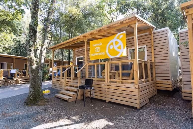 Stacaravan met een overdekte houten veranda, een barbecue en een gele Eurocamp-vlag, langs een pad onder de bomen