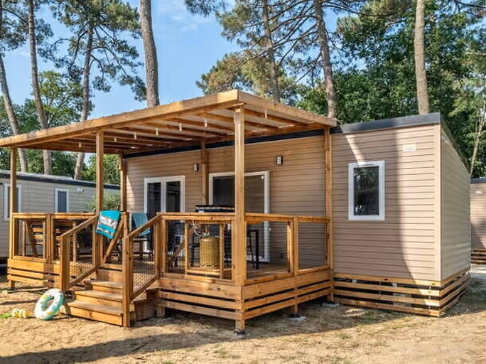 Beige stacaravan met een houten terras onder een pergola, een groene zwemband tegen de trap en dennen erachter