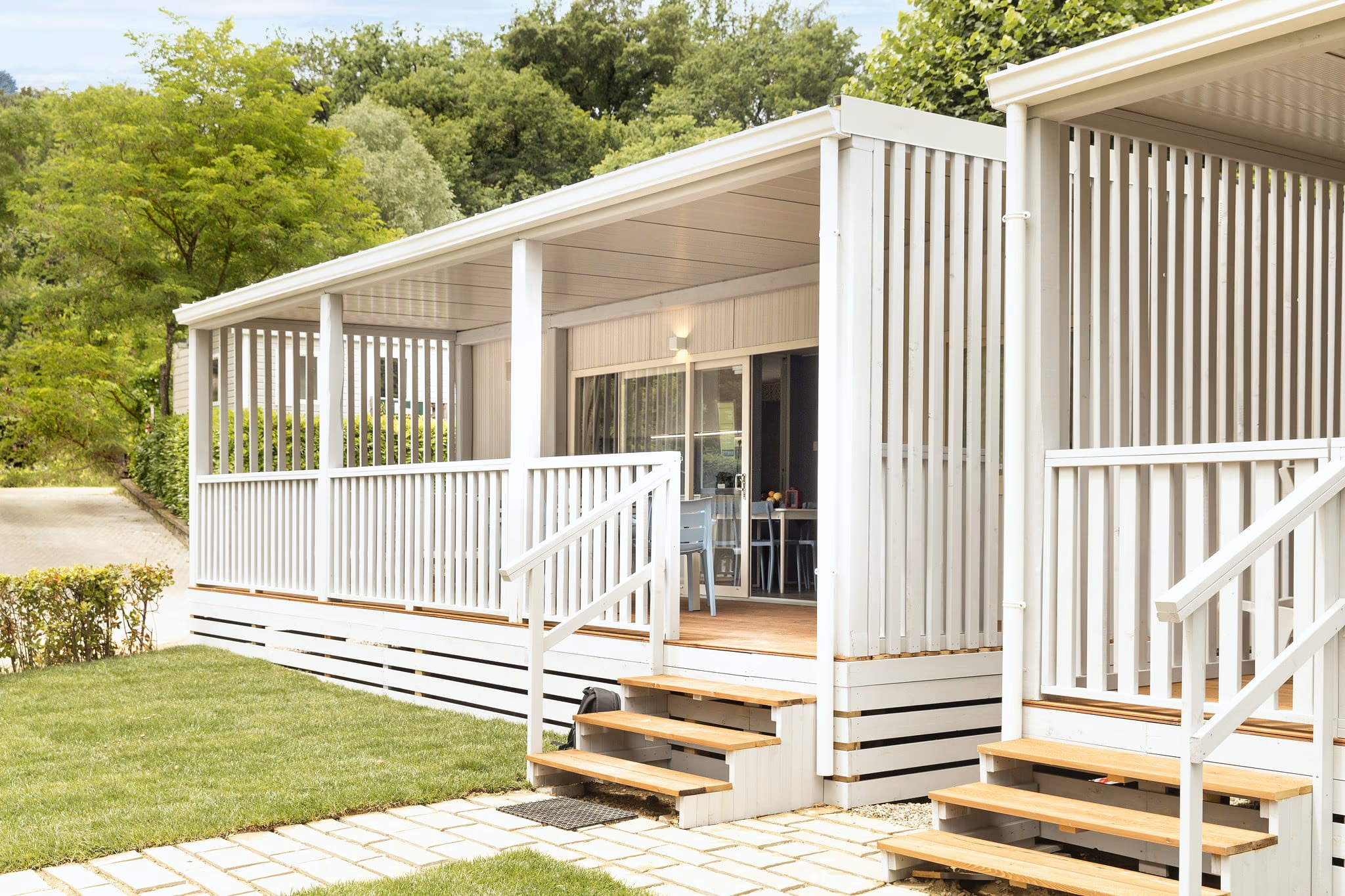 Twee witte stacaravans met een lang overdekt terras en houten traptreden op Camping Bella Italia, met bomen erachter