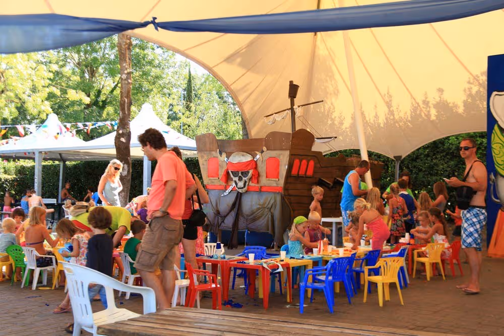Kinderen en begeleiders aan tafels onder de tent van de miniclub op Camping Bella Italia