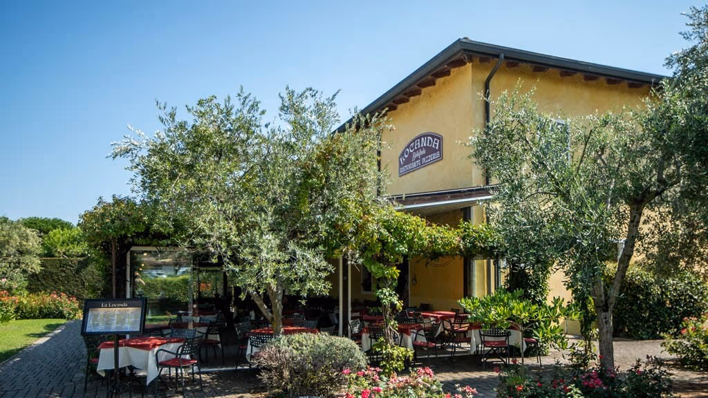 Restaurantgebouw met terras tussen olijfbomen op Camping Bella Italia aan het Gardameer