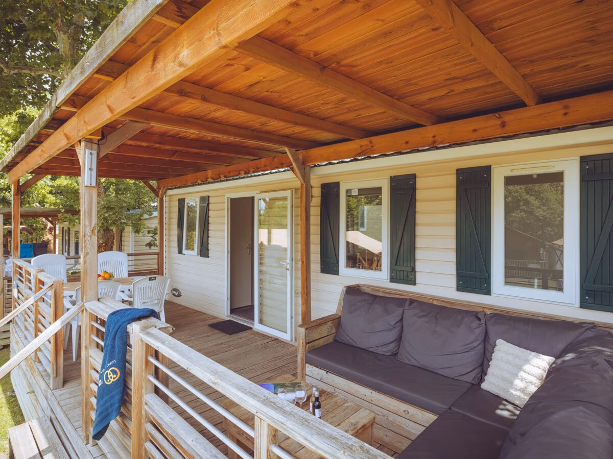 Veranda onder een houten balkenkap met een grote loungebank en witte stoelen bij een stacaravan met groene luiken op Camping Bella Italia