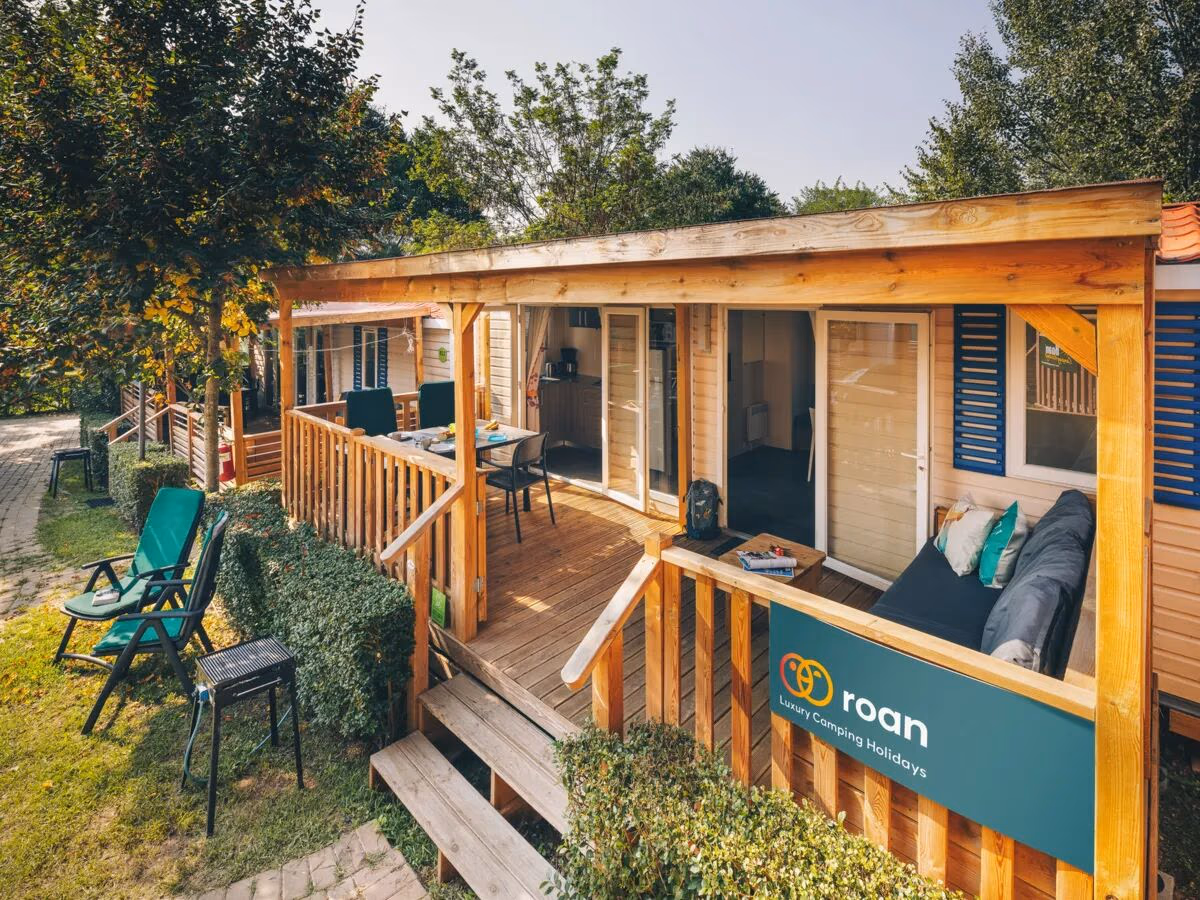 Stacaravan met een houten veranda met eettafel en loungebank op Camping Bella Italia, met groene klapstoelen in het gras ernaast