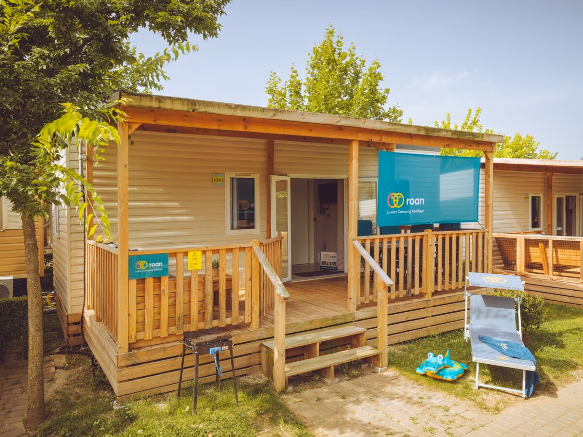 Stacaravan met een houten overkapping en trap naar de veranda op Camping Bella Italia, met een kinderstoeltje en speelgoed in het gras