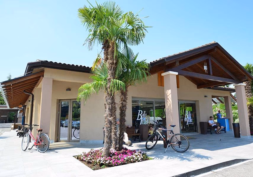 Winkelgebouw van Camping Bella Italia met palmbomen, een bloemperk en geparkeerde fietsen voor de deur