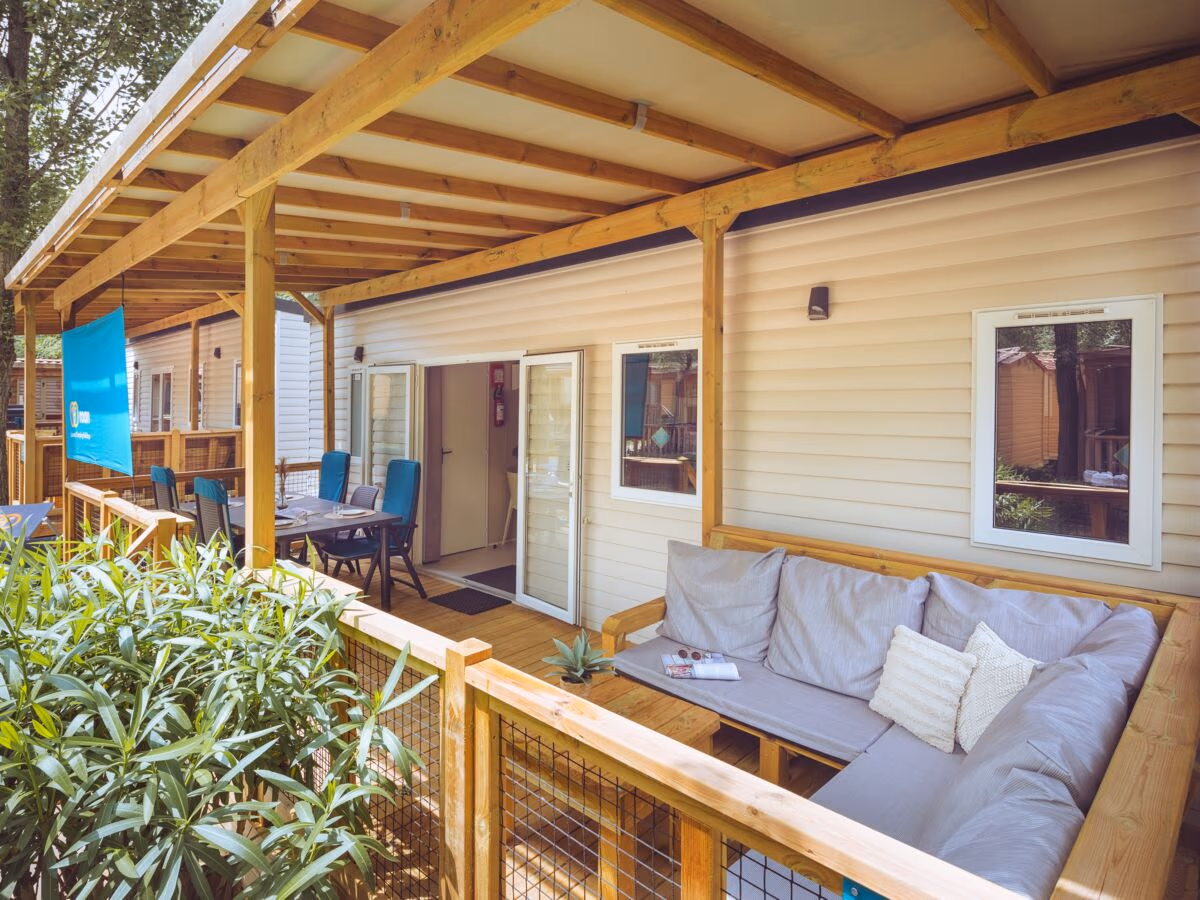 Overdekte veranda met een grijze hoekbank en een eettafel met blauwe stoelen bij een stacaravan