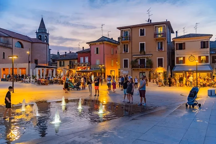 Een plein in Bardolino bij valavond met waterstraaltjes in de bestrating, terrassen en kleurrijke huizen